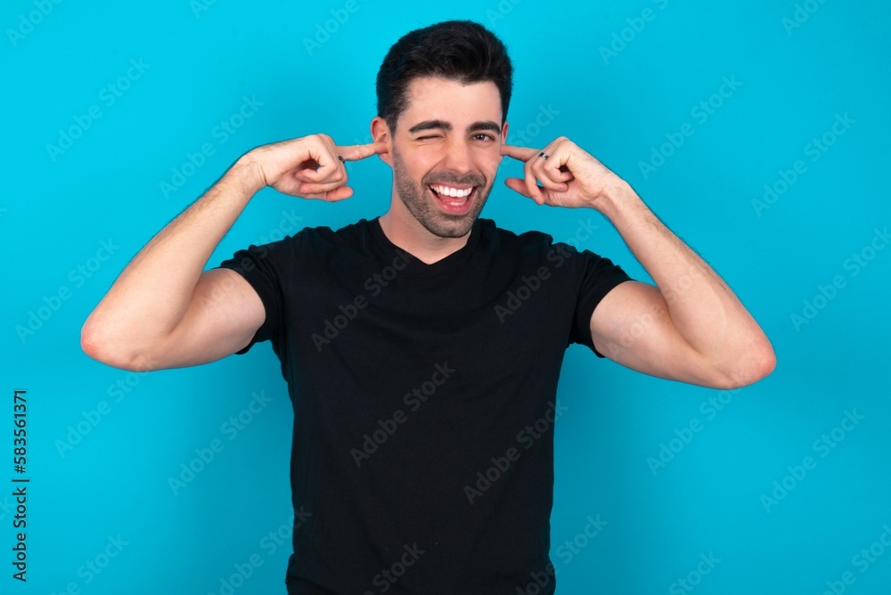 Happy Young man wearing black T-shirt over blue studio background ignores loud music and plugs ears with fingers asks to turn off sound Happy Young man wearing black T-shirt over blue studio background ignores loud music and plugs ears with fingers asks to turn off sound