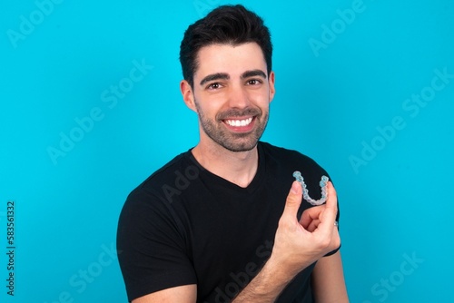 Young man wearing black T-shirt over blue studio background holding an invisible aligner ready to use it. Dental healthcare and confidence concept.