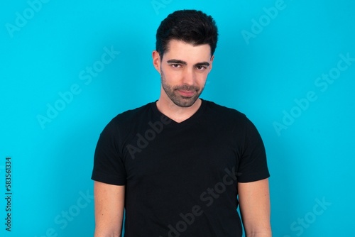 Young man wearing black T-shirt over blue studio background with nice beaming smile pleased expression. Positive emotions concept