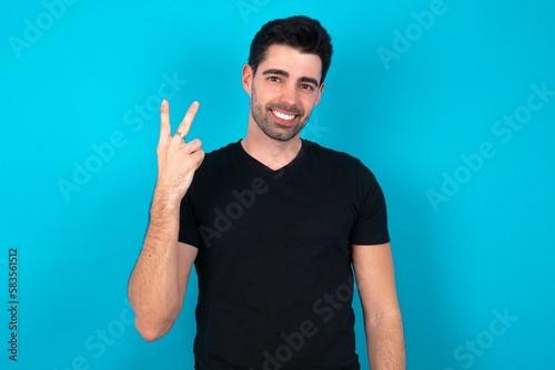 Young man wearing black T-shirt over blue studio background smiling and looking friendly, showing number two or second with hand forward, counting down © Jihan