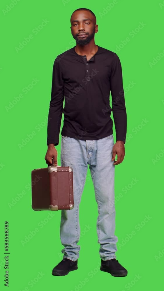 Vertical video: Front view of casual person holding suitcase luggage on camera and waiting for something, standing over green screen backdrop. Young man with briefcase carrying baggage in studio Vertical video: Front view of casual person holding suitcase luggage on camera and waiting for something, standing over green screen backdrop. Young man with briefcase carrying baggage in studio