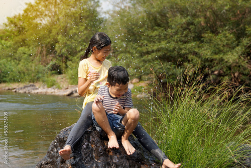 Asian girls and boys take a break from playing in the river to sit on the rocks. Both of them laughed and dodged the water that had been poured over them. Water droplets spread and look fresh.