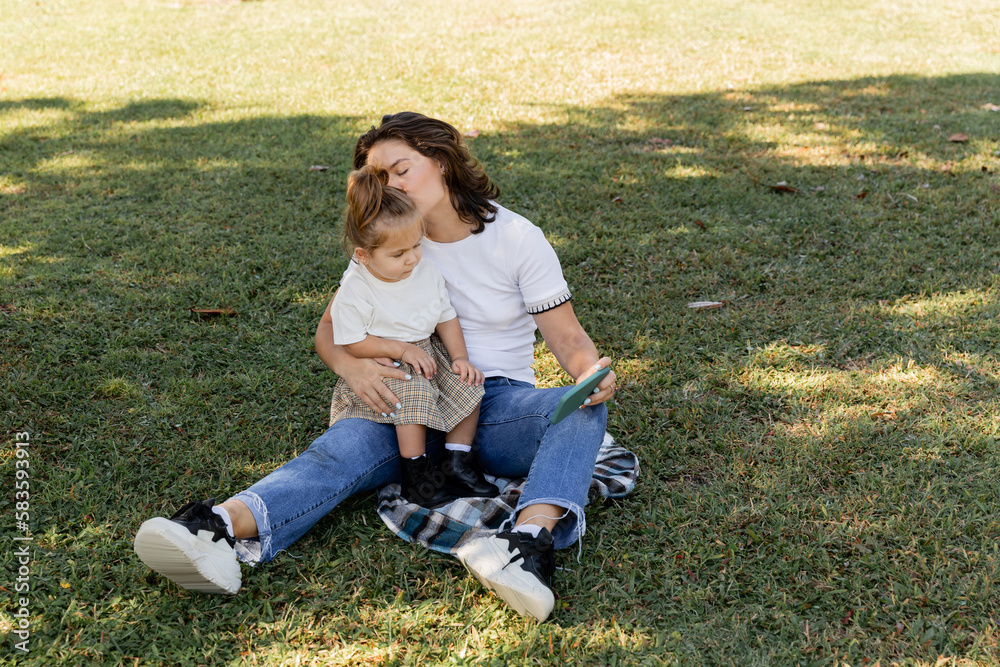 mother kissing baby girl while holding smartphone and sitting on lawn.