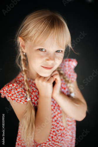 Portrait of an 8-year-old blonde girl with two pigtails, in a summer floral dress on a dark background, smiling and looking at the camera.