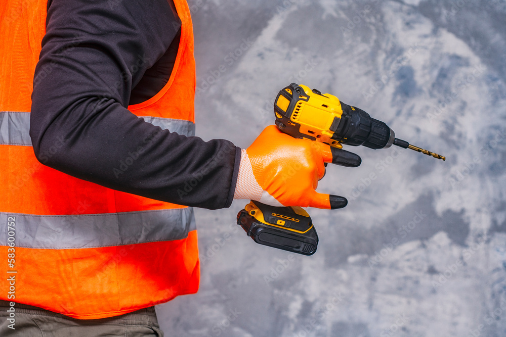 Male worker holds a close-up electric cordless screwdriver in his hands against the background of a construction tool and a concrete wall. Male worker holds a close-up electric cordless screwdriver in his hands against the background of a construction tool and a concrete wall.