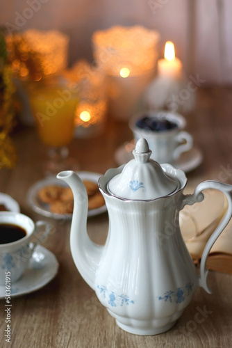 Cup of tea or coffee, plate of cookies, cup of blueberries, plate of chocolate, glass of juice, book, reading glasses, teapot, flowers and lit candles on the table. Brunch or afternoon tea concept. 