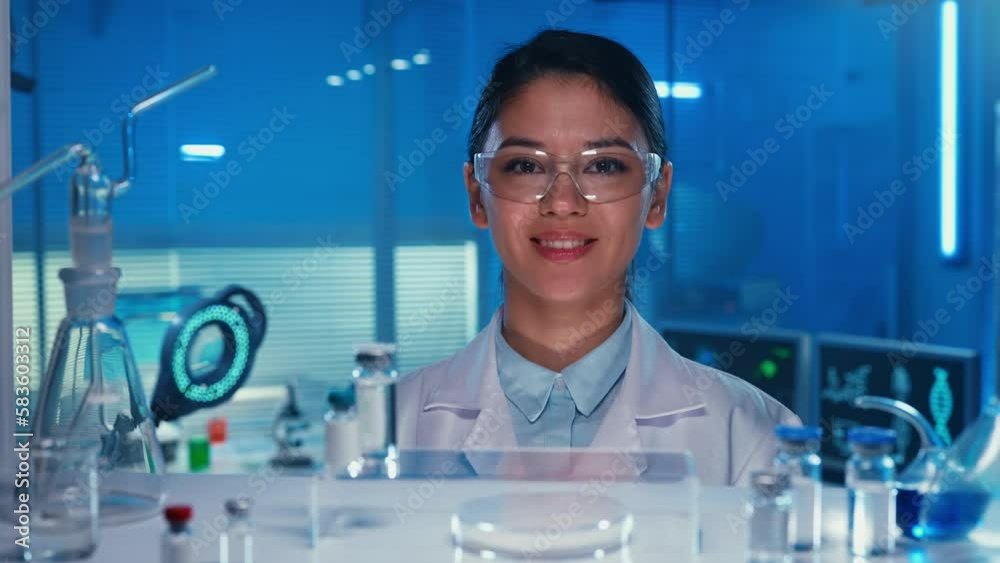 Portrait of an Asian female doctor looking at the camera and smiling. Female laboratory assistant in glasses and a white medical coat in a laboratory or hospital with blue light. Close up.