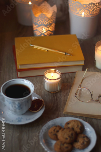 Cup of tea or coffee  plate of cookies  books  e-reader  pencil and lit candles on the table. Selective focus.