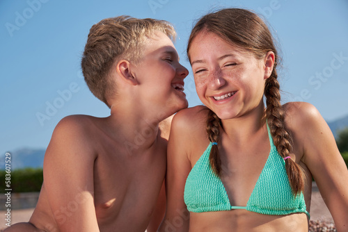Happy joyful boy and girl are whispering while sitting on the seashore. Sunny summer day, vacation