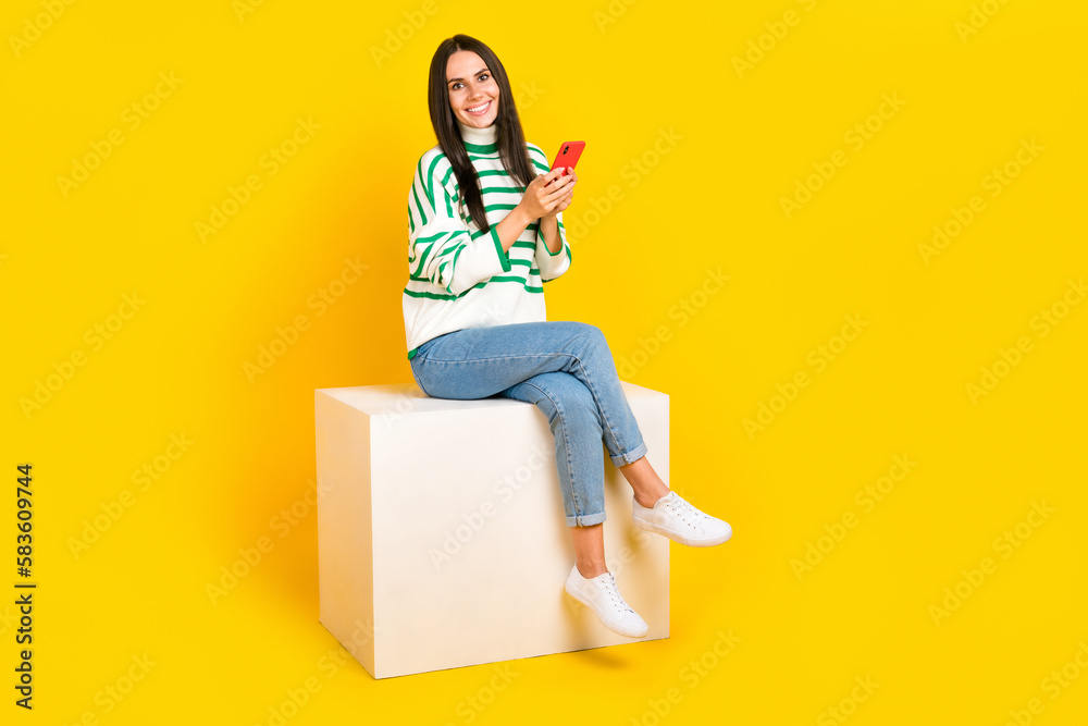 Full length photo of adorable positive lady wear stylish striped clothes use samsung iphone empty space isolated on yellow color background Full length photo of adorable positive lady wear stylish striped clothes use samsung iphone empty space isolated on yellow color background