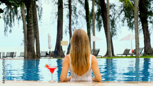 Young Woman with glass of cocktail in White Bikini in Swimming Pool, Tanned on Summer Vacation. Relaxing Concept at Spa Resort. Girl Enjoy Beach Holidays at Luxurious Beachfront Hotel. Back view © TravelMedia