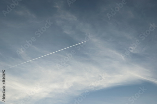 Airplane trail in blue cloudy sky background. Horizontal linear trace from a flying fast aircraft in the distance
