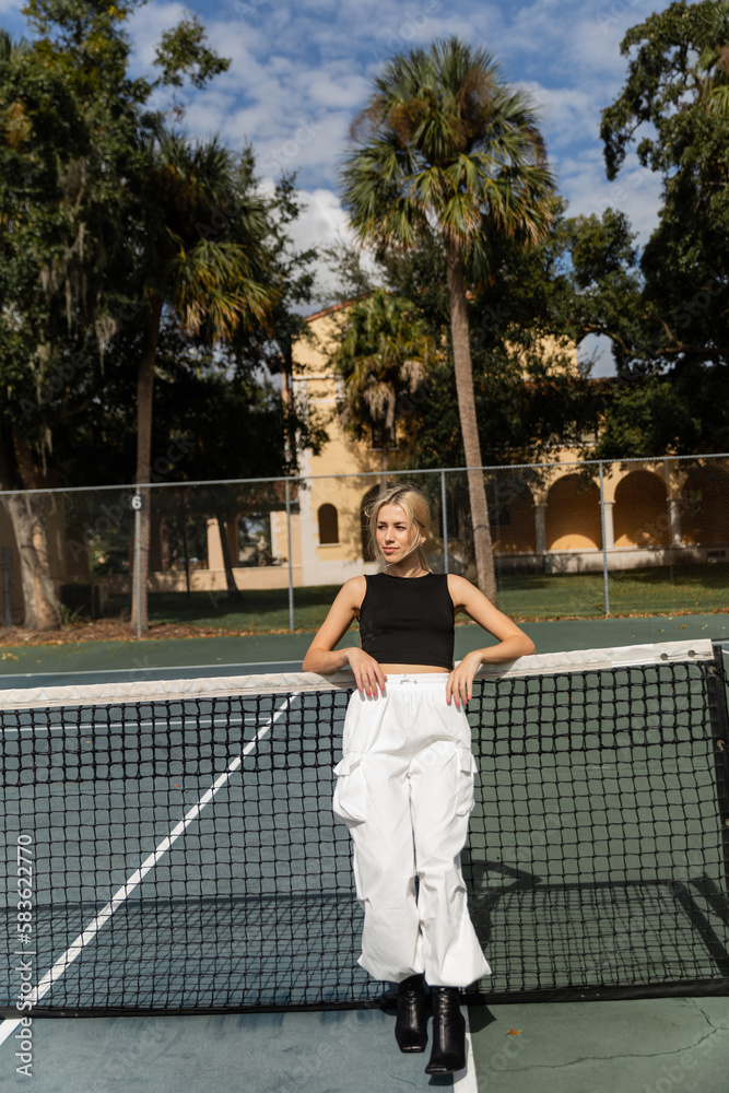 full length of blonde woman in tank top and white cargo pants leaning on net in tennis court.