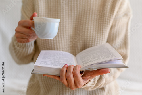 A woman in a white jumper is holding a book and a cup of white coffee.