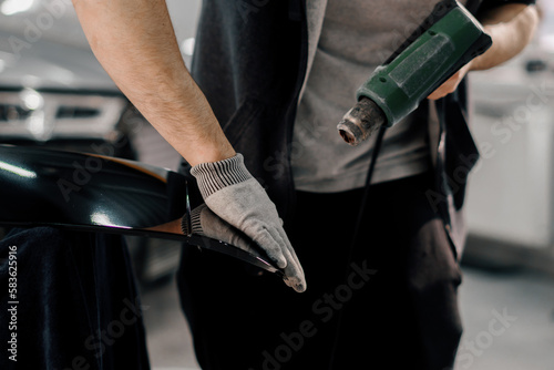 A worker covers a car spoiler with a film in the workshop using a hair dryer and a spatula close-up Car detailing and care