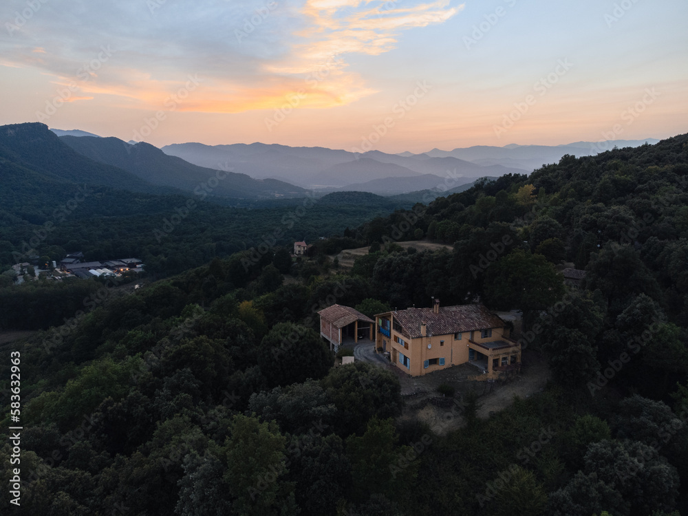 Aerial photograph of a beautiful house in the middle of nature, surrounded by a lush forest and majestic mountains during sunset. The image shows a spectacular view of the house.