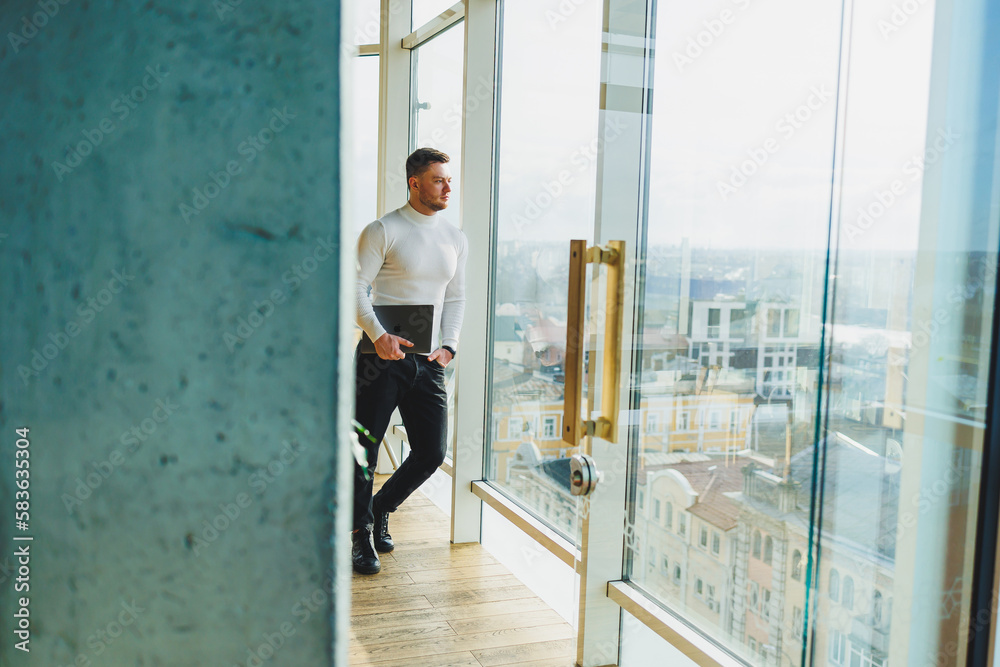 Fototapeta premium A young man in a modern workspace stands near a large window. A man in casual clothes with a laptop in his hands. Remote work. Nice young manager