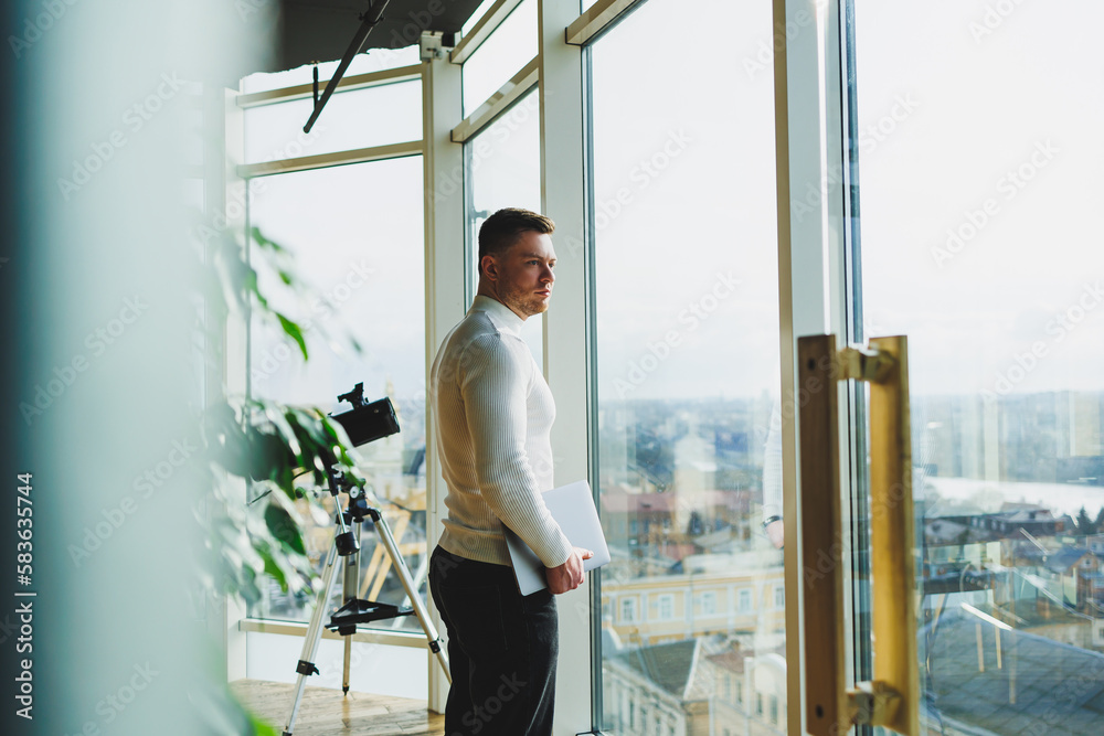 A young man in a modern workspace stands near a large window. A man in casual clothes with a laptop in his hands. Remote work. Nice young manager A young man in a modern workspace stands near a large window. A man in casual clothes with a laptop in his hands. Remote work. Nice young manager