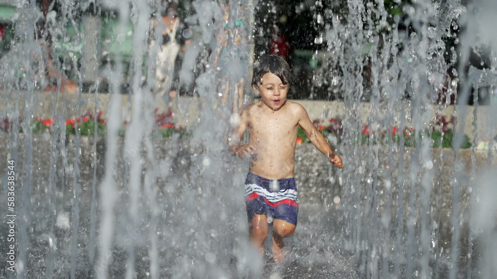 Little boy running at water jets in city park during summer day. Small child runs in refreshing water splashing in slow motion Little boy running at water jets in city park during summer day. Small child runs in refreshing water splashing in slow motion