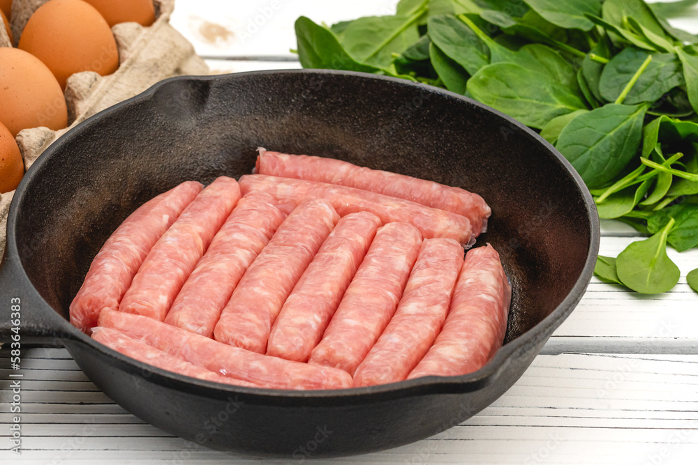 Raw chicken breakfast sausages on frying pan, eggs, and spinach. Ingredients for breakfast close up on white wooden board Raw chicken breakfast sausages on frying pan, eggs, and spinach. Ingredients for breakfast close up on white wooden board
