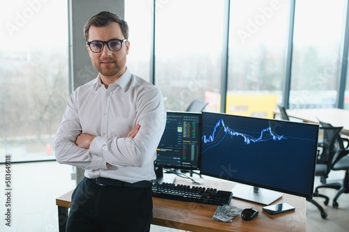 Man trader in formalwear sitting at desk in frot of monitors with charts and data at office browsing laptop checking documents analyzing stocks price changes concentrated. photo