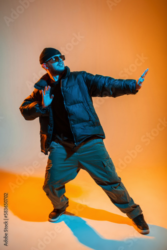 Stylish trendy handsome professional hip hop dancer man with sunglasses and cap in fashion black clothes with winter down jacket and sneakers dances in creative studio with orange and cold neon light