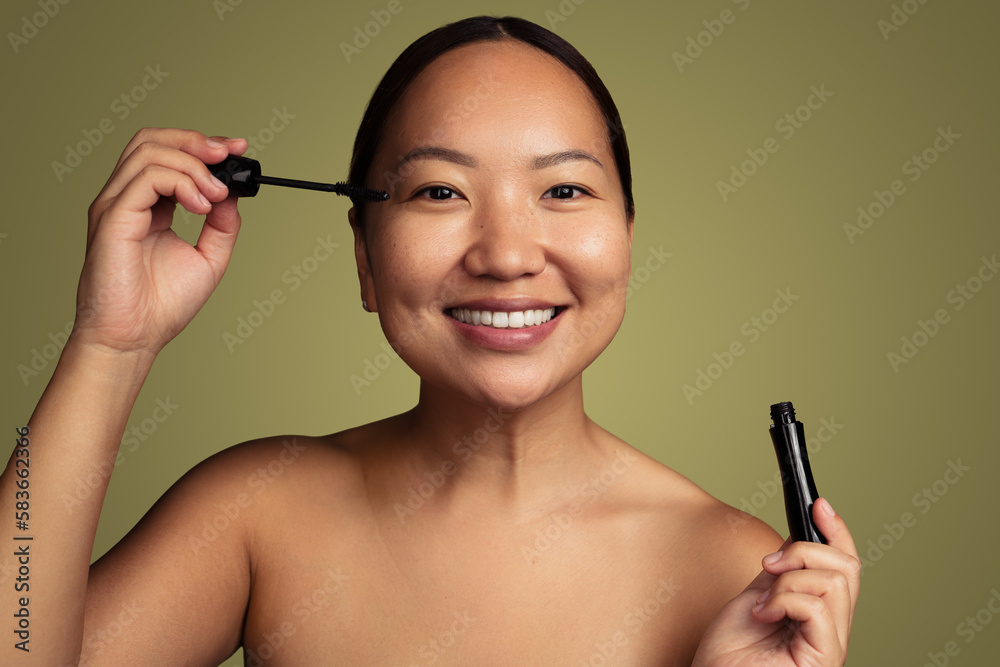 Fototapeta premium Joyful young Asian woman applying mascara in green studio