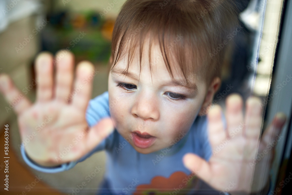 Cute little child, blond toddler boy sitting indoors looking through window