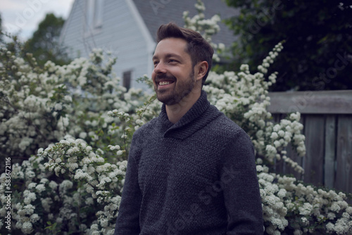 Portrait of a 35 year old caucasian man standing outside wearing a wooden sweater. He is standing in front of a white lilac tree and a blue house.