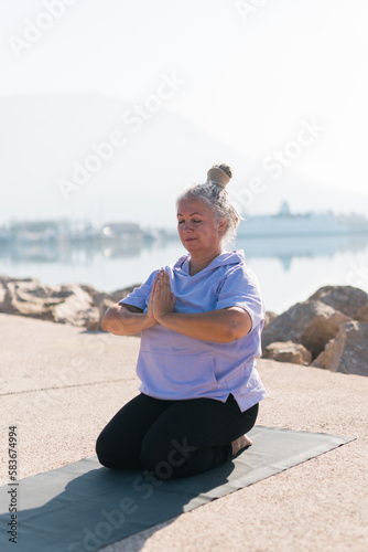 Senior woman with dreadlocks in stretching position by the sea at morning. Elderly woman doing yoga near beach.