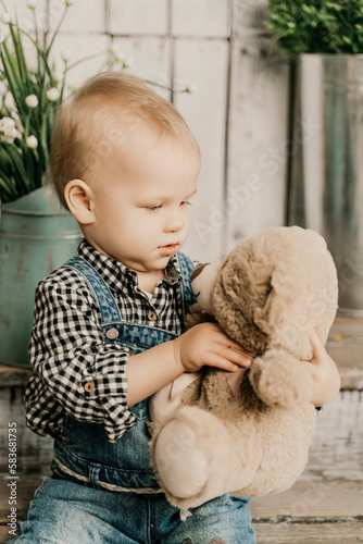 Girl 1 year old. sits and holds a teddy bear, she is dressed in a plaid shirt and denim overalls on a wooden background with green potted plants. Fun moments with kids.