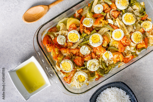 Gomes de sa codfish roasted in olive oil with tomatoes, peppers, onions, boiled eggs and oregano. In a rectangular glass baking dish. Detail wooden spoon and olive oil.