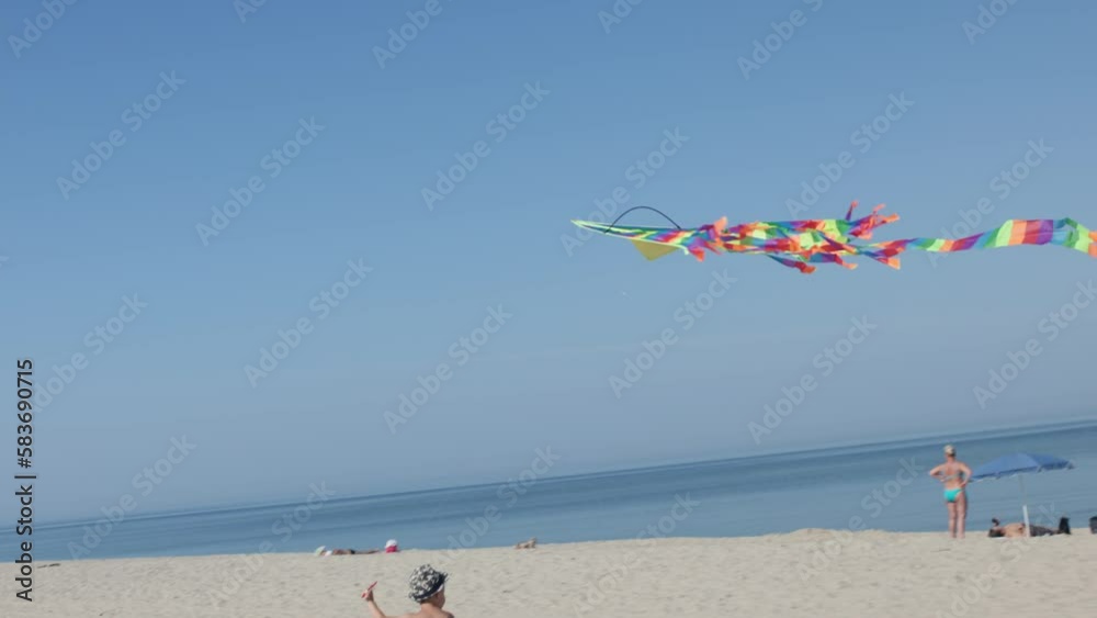 little kid boy running on beach trying to fly colorful kite in sky. bright striped kite flying in blue sky kid holding string run on white sand seaside near sea ocean edge. children summer activity 