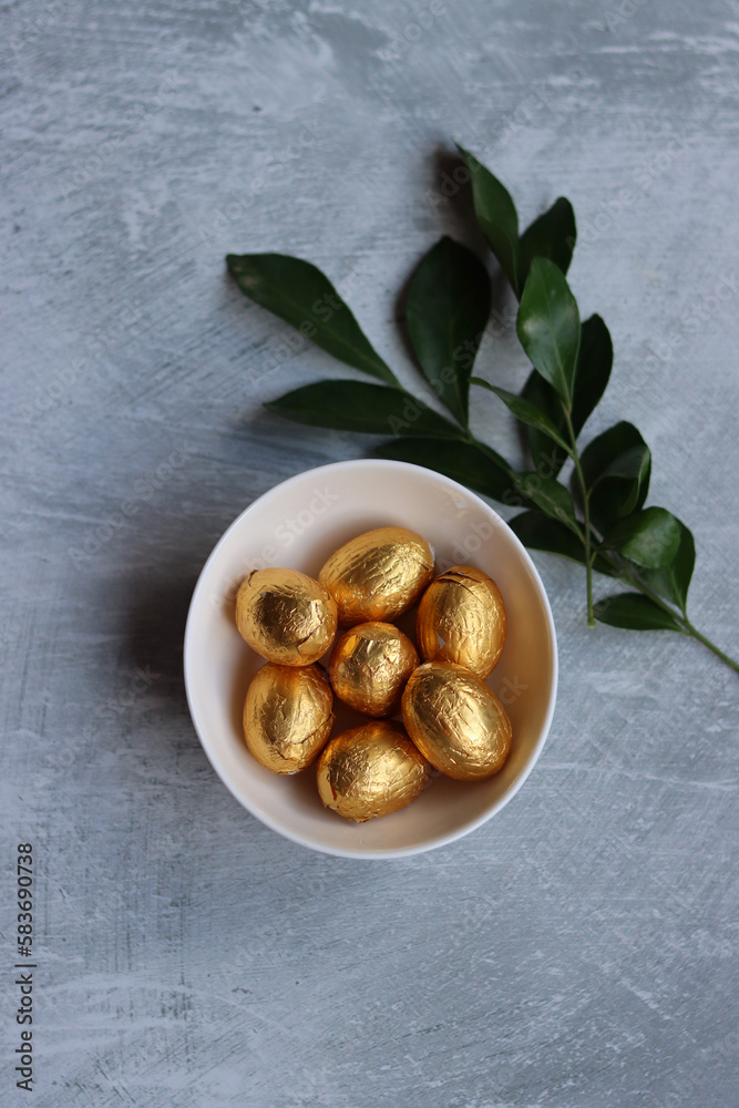 Obraz premium Chocolate Easter Egg Wrapped in Gold Foil. White spring flowers and Ester sweets top view photo. Bright spring still life. 