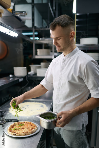 Male chef sprinkling fresh greenery over traditionally made pizza