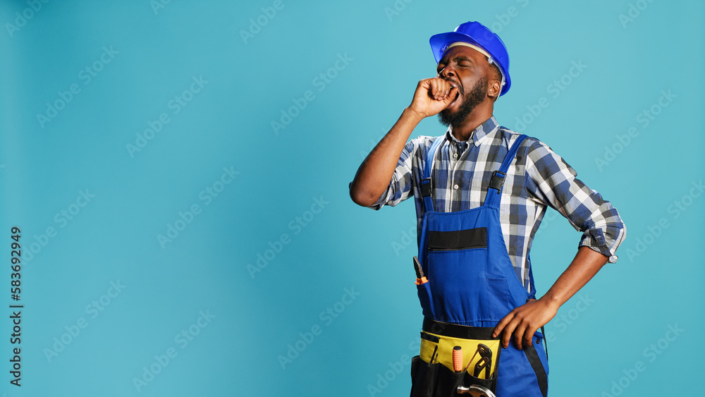 Male builder yawning on camera feeling worn out, man dressed in overalls and hardhat falling asleep in studio. Tired handyman feeling exhausted and drained after construction project.