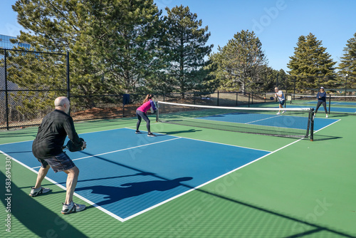 A competitive doubles game of pickleball with a group of men and women on a blue and green court in spring.