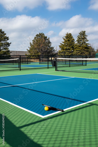 View of a pickleball complex with a paddle and yellow ball on blue and green courts beside a playground in a suburban park in early spring. © pics721