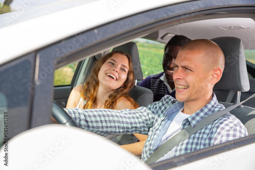 Cheerful young man driving car with laughing friends in passenger seats. Friendly road trip and travel concept © JackF