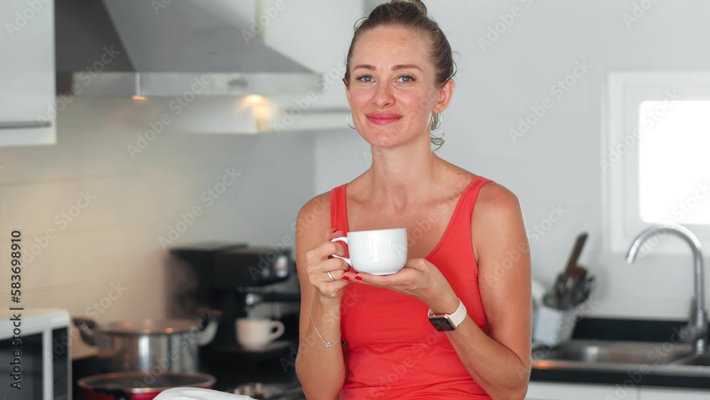 Woman drinking tea from cup on kitchen during cooking meal and looking in camera Woman drinking tea from cup on kitchen during cooking meal and looking in camera