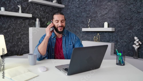 a young guy works at home at the computer talks on the phone took a break makes an appointment by phone. View of camera sliding left to smiling happy freelancer in a denim shirt sitting at the table