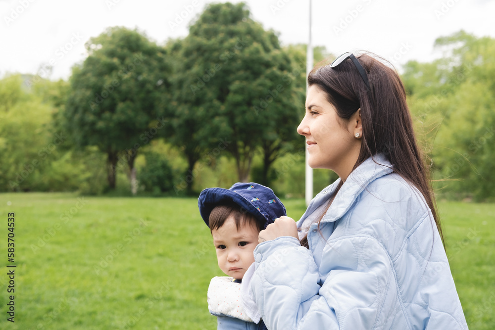 A mother carrying her male infant with a baby carrier while walking at the park in spring season and wearing blue clothes and sun glasses. Biracial and multicultural family. A mother carrying her male infant with a baby carrier while walking at the park in spring season and wearing blue clothes and sun glasses. Biracial and multicultural family.