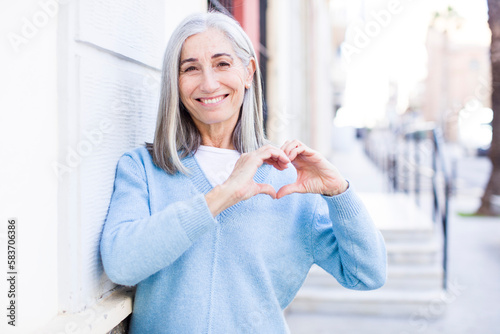 senior retired pretty white hair woman smiling and feeling happy, cute, romantic and in love, making heart shape with both hands