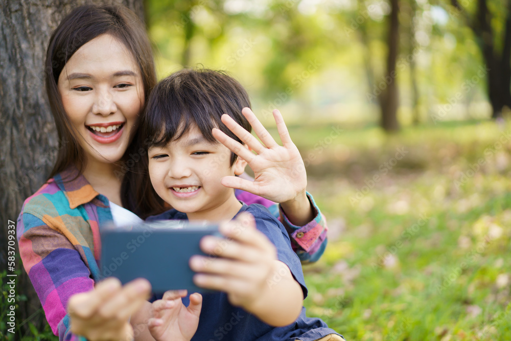 Happy cheerful Asian little boy using smartphone to take a selfie photography with his mother at the park in morning. Happy family time concept, woman staying with her little son in weekend. Happy cheerful Asian little boy using smartphone to take a selfie photography with his mother at the park in morning. Happy family time concept, woman staying with her little son in weekend.
