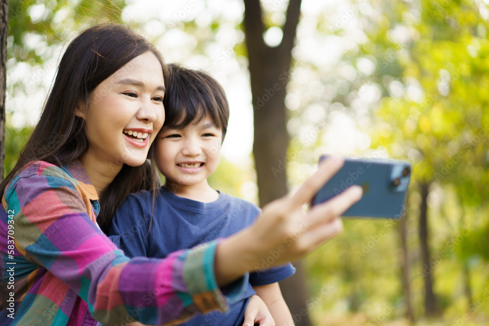 Fototapeta premium Happy cheerful Asian little boy using smartphone to take a selfie photography with his mother at the park in morning. Happy family time concept, woman staying with her little son in weekend.