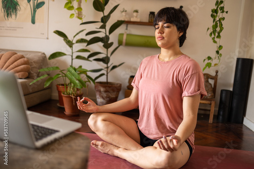 Young latin woman with a short hair meditating with her eyes closed. She is wearing pink shirt and sitting in the room full of green plants.