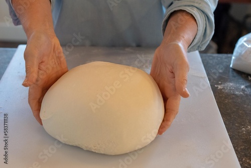 A large bun of dough rises on a white plastic board with a red glass bowl in the background. The large ball of dough is made with white flour. There's a window in the background with natural light.