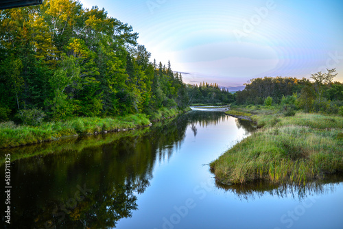 A scenic meandering river as the sun sets in the distance. The trees and lush green reeds are reflected in the smooth water. The serene lake has lush riverbanks with green grass and a thick forest.