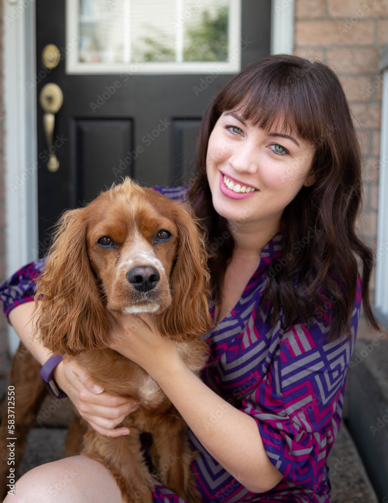 Outdoor portrait of a caucasian woman hugging her cocker spaniel dog sitting outside on her front step of her town house. The lady is smiling and she has brown hair and blue eyes. 