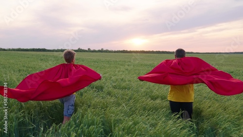 Happy boys play superheroes they run across green field in red cloak  cloak flutters in wind. Childrens games and dreams. Slow motion. teenager dreams of becoming superhero. Young boys in red cloak